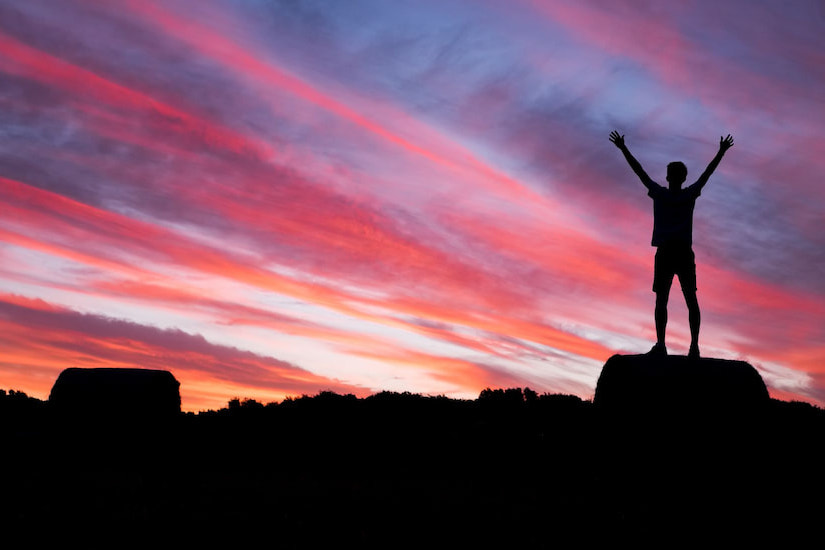 young person standing on a rock with their arms up looking into the sunset. life after cancer image