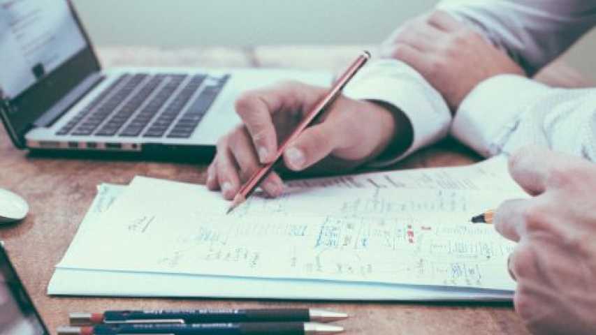 Image of hands writing on a paper next to a laptop 